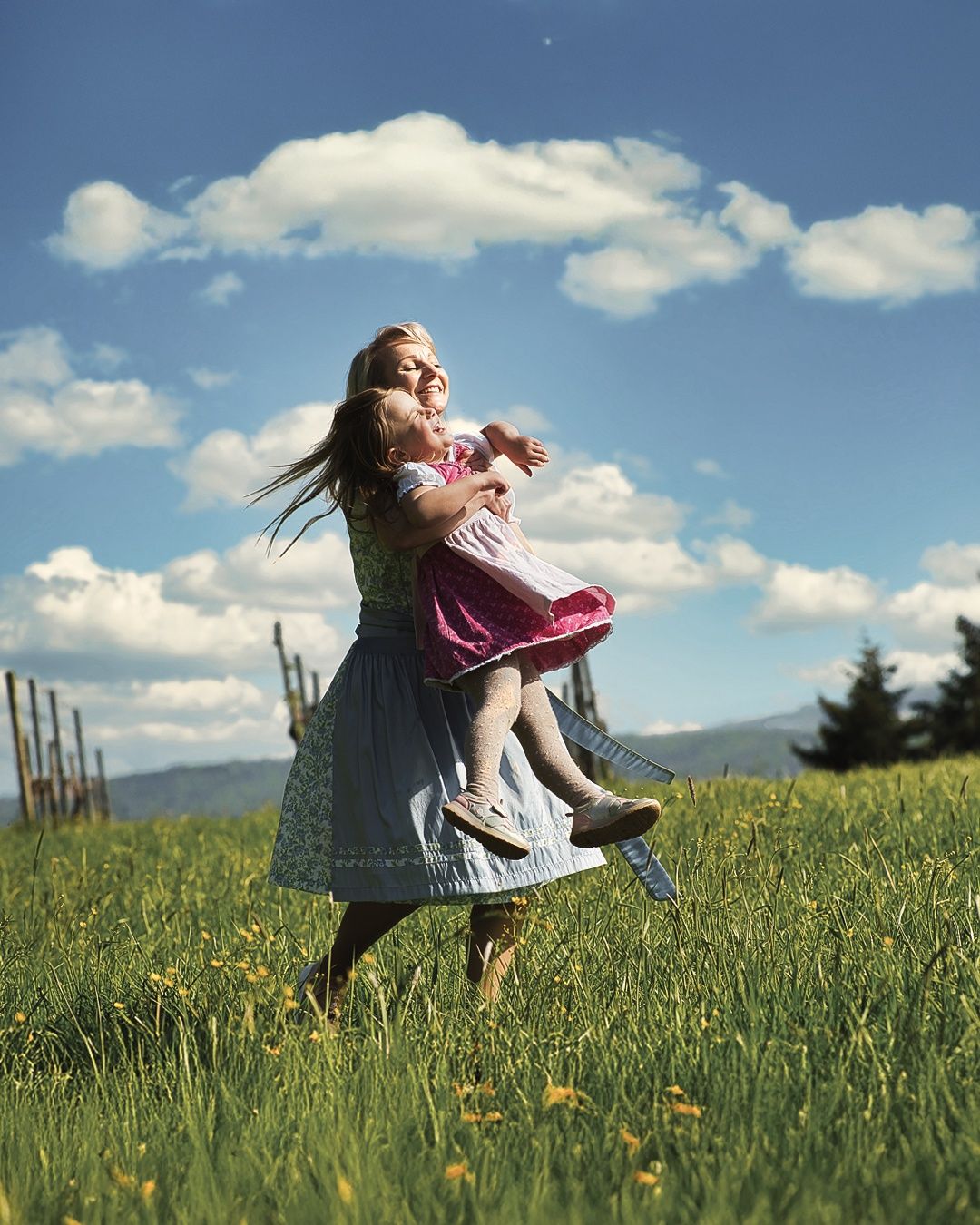 Eine Frau hält fröhlich ein kleines Mädchen in einem rosa Kleid, beide lächeln in einem üppigen, grünen Feld unter einem blauen Himmel mit verstreuten Wolken.