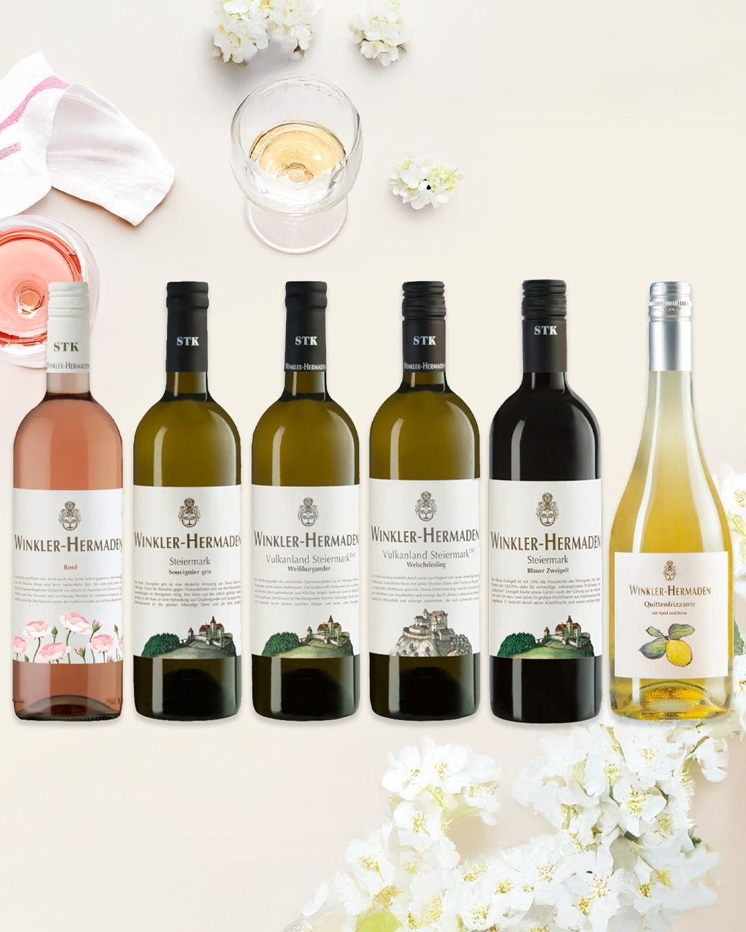 Sieben Flaschen Winkler-Hermaden-Wein in verschiedenen Farben von Rosé bis Gelb, arrangiert mit einem Glas Wein und einem Tuch. Der Hintergrund zeigt ein weißes Blumenmuster.