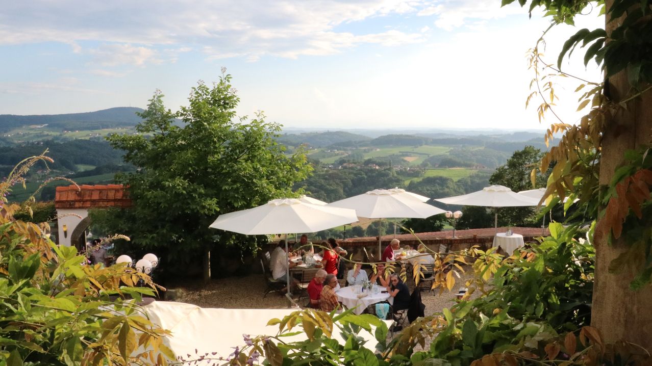 Eine Gruppe von Menschen isst unter weißen Sonnenschirmen auf einer Terrasse mit einer malerischen Aussicht auf das Tal und die Hügel unter einem bewölkten Himmel.