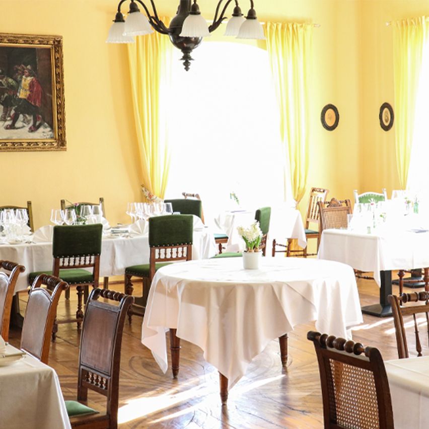 Ein Restaurant mit gelben Wänden und Holzböden. Tische mit weißen Tischdecken, einige mit Blumen in Vasen. Stühle sind um die Tische herum angeordnet. Eine Kronleuchter hängt von der Decke. Ein Gemälde hängt an der Wand.