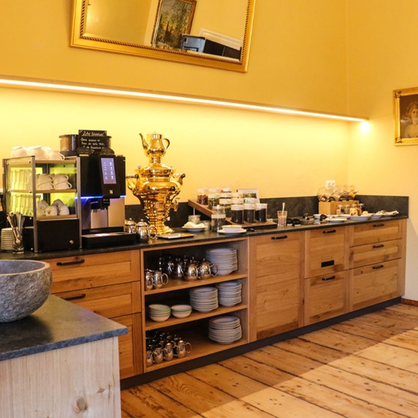 Eine Küche mit Holztheken, Regalen und einer großen Schüssel links. Eine Kaffeemaschine, ein goldener Teepott und ein Bilderrahmen stehen auf der Theke. Es gibt auch andere Gegenstände wie eine Schüssel, eine Tasse und einen Teller.