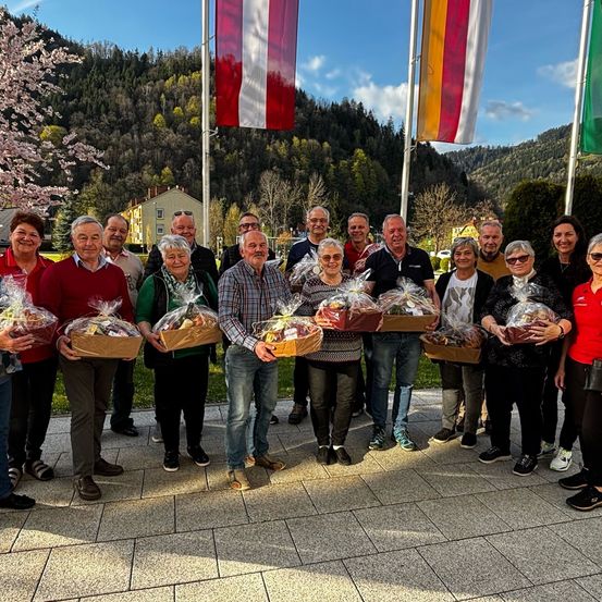 Eine Gruppe von Menschen, die Geschenkkörbe halten, steht auf einer Betonfläche. Hinter ihnen befinden sich Fahnen, ein Haus und ein Berg.