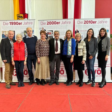 Eine Gruppe von Menschen steht auf einem roten Teppich vor drei Bannern. Die Banner repräsentieren die Jahre 1990, 2000 und 2010. Sie scheinen ein Jubiläum zu feiern.