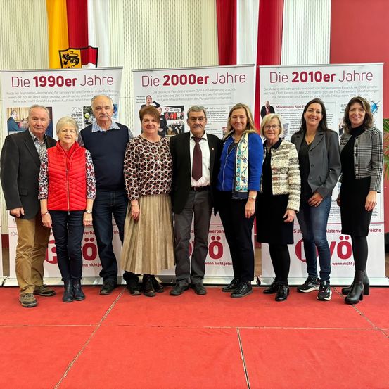 Eine Gruppe von Menschen steht auf einem roten Teppich vor drei Bannern. Die Banner repräsentieren die Jahre 1990, 2000 und 2010. Sie scheinen ein Jubiläum zu feiern.