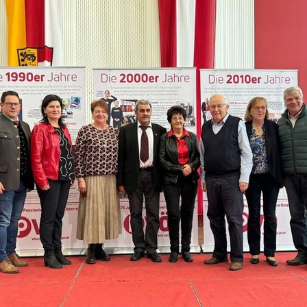 Gruppe von Menschen in formeller und lässiger Kleidung, die auf einem roten Teppich stehen, mit drei Plakaten im Hintergrund. Die Plakate zeigen verschiedene Jahre, darunter 1990, 2000 und 2010.