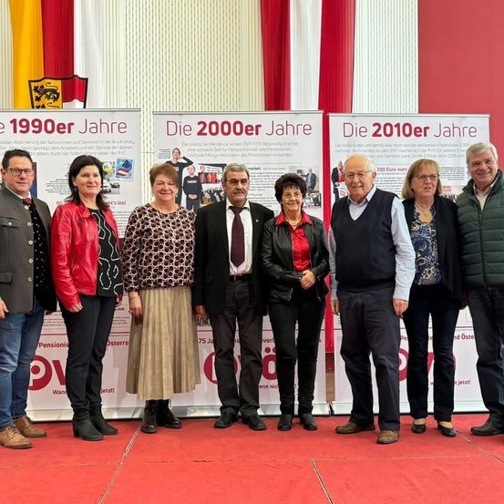 Gruppe von Menschen in formeller und lässiger Kleidung, die auf einem roten Teppich stehen, mit drei Plakaten im Hintergrund. Die Plakate zeigen verschiedene Jahre, darunter 1990, 2000 und 2010.