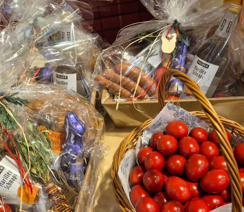 Mehrere Körbe mit verschiedenen Artikeln wie Brot, Schokolade und Tomaten sind auf einem Tisch angeordnet. Jeder Korb hat eine eigene Plastikabdeckung und dekorative Elemente.