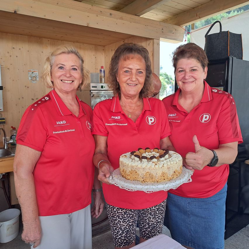 Drei Frauen in roten Uniformen lächeln und posieren mit einem Kuchen auf einem Teller. Die Frau auf der linken Seite hält ein Kuchenmesser. Hinter ihnen ist eine Holzwand mit einem Waschbecken und einem Kühlschrank.