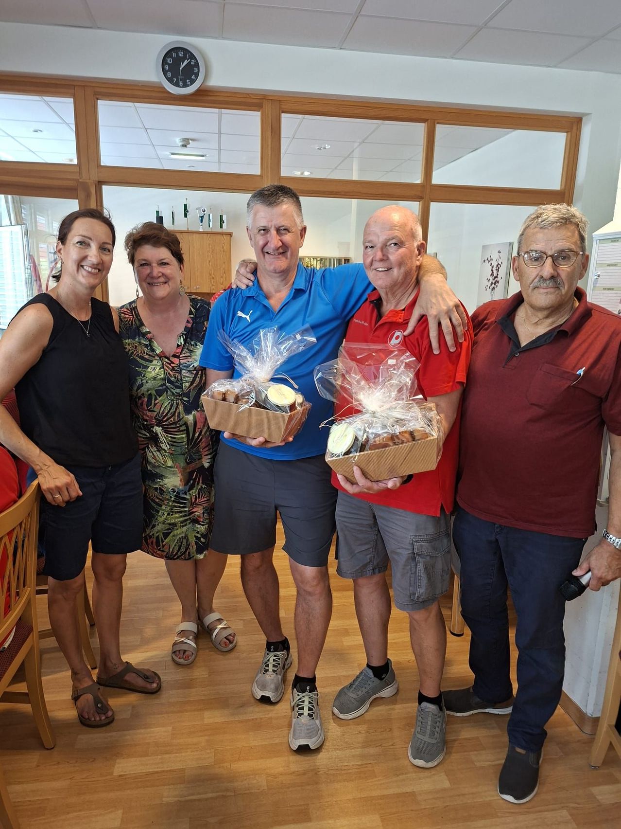 Five people are posing for a photo. Two men are holding boxes of food. One is in a blue shirt and the other in a red shirt. Three women stand next to the men, smiling. They are in a room with wooden floors and glass windows.