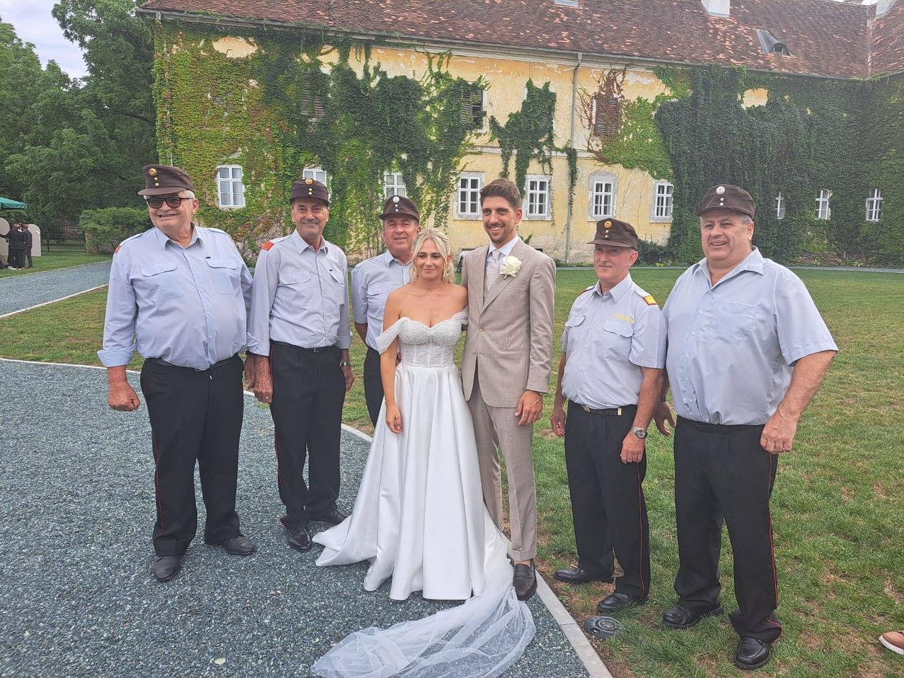 Eine Gruppe uniformierter Männer und eine Frau in einem Hochzeitskleid stehen vor einem Gebäude mit Efeu.