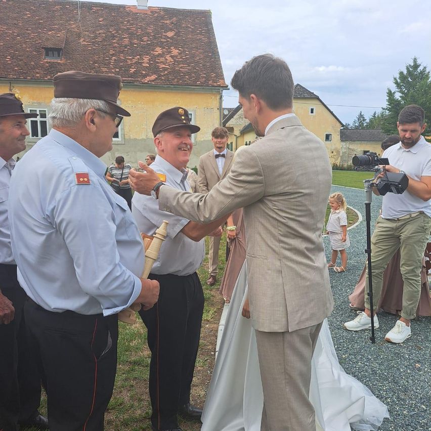 Eine Hochzeitszeremonie im Freien, mehrere Personen anwesend, einer hält eine Pfeife, ein Bräutigam in einem hellen Anzug und ein Fotograf, der den Moment einfängt.