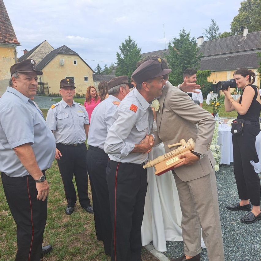 Eine Gruppe uniformierter Männer steht auf einem Rasen mit einem Mann in Anzug, der eine Trophäe hält. Hinter ihnen macht eine Frau ein Foto. Ein Tisch mit Blumen und einer Frau in einem schwarzen Oberteil ist im Hintergrund.