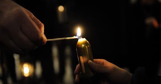 Eine Person zündet eine Kerze mit einem Streichholz an. Die Kerze wird in einer Hand gehalten, während das Streichholz in der anderen gehalten wird. Die Szene ist schwach beleuchtet.