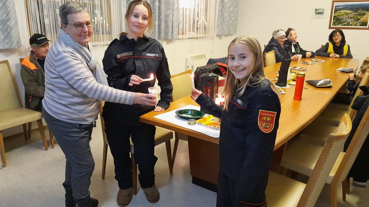 Drei Personen in Uniform, zwei mit brennenden Kerzen, stehen neben einem Tisch mit einer Schüssel und einer Laterne. Hinter ihnen sitzt eine Person auf einem Stuhl.