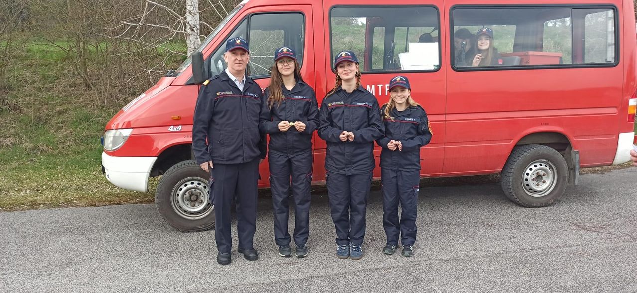Fünf Personen in Uniformen stehen vor einem roten Fahrzeug und halten Abzeichen. Sie scheinen Feuerwehrleute zu sein.