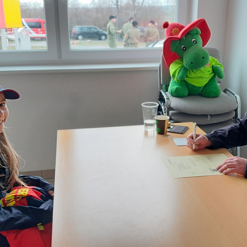 Ein junges Mädchen sitzt an einem Tisch mit einem Spielzeugalligator mit rotem Hut. Ein Mann in Uniform unterschreibt ein Papier. Ein Fenster zeigt Autos und Menschen draußen.