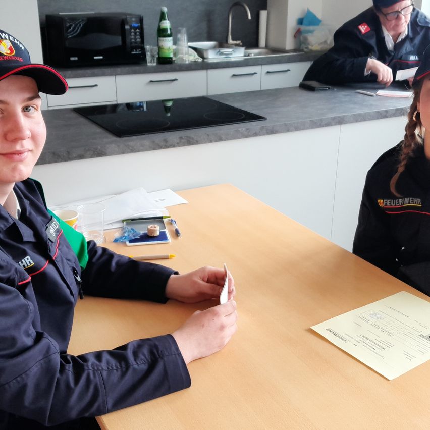 Zwei Feuerwehrleute sitzen an einem Tisch, einer hält ein Stück Papier in der Hand. Eine Mikrowelle, eine Flasche und ein Becher stehen auf der Arbeitsplatte. Ein weiterer sitzt hinter ihnen.