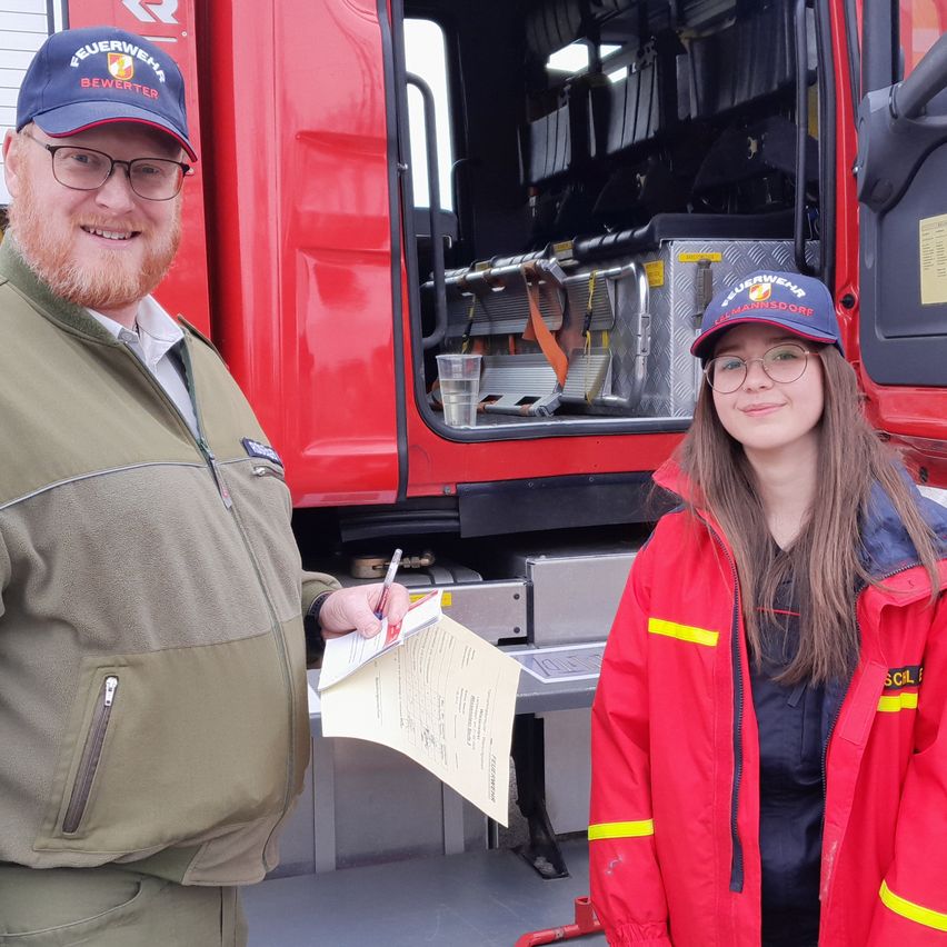 Zwei Feuerwehrleute stehen vor einem roten Feuerwehrauto, einer hält ein Papier und einen Stift. Die Frau lächelt und beide tragen reflektierende Westen.