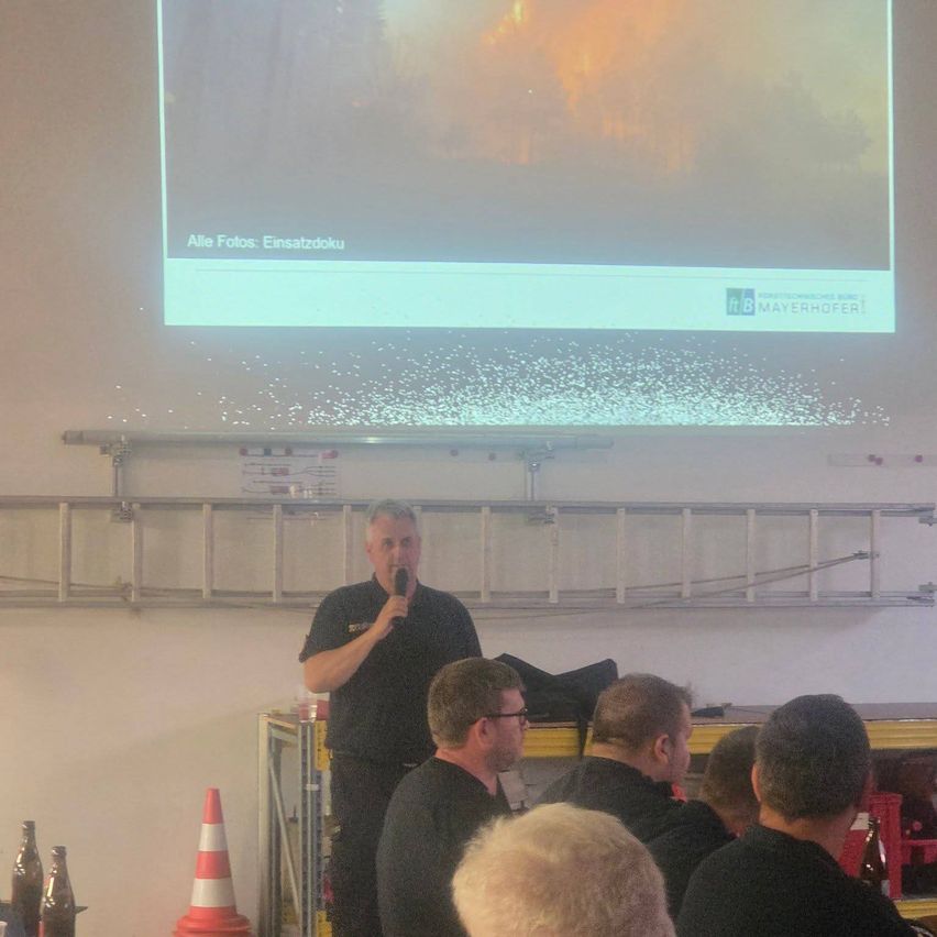 Ein Mann steht vor einer Projektionsleinwand mit einem Feuerbild und spricht in ein Mikrofon. Ein Publikum von Männern sitzt vor ihm. Es gibt Leitern und Flaschen auf dem Tisch.