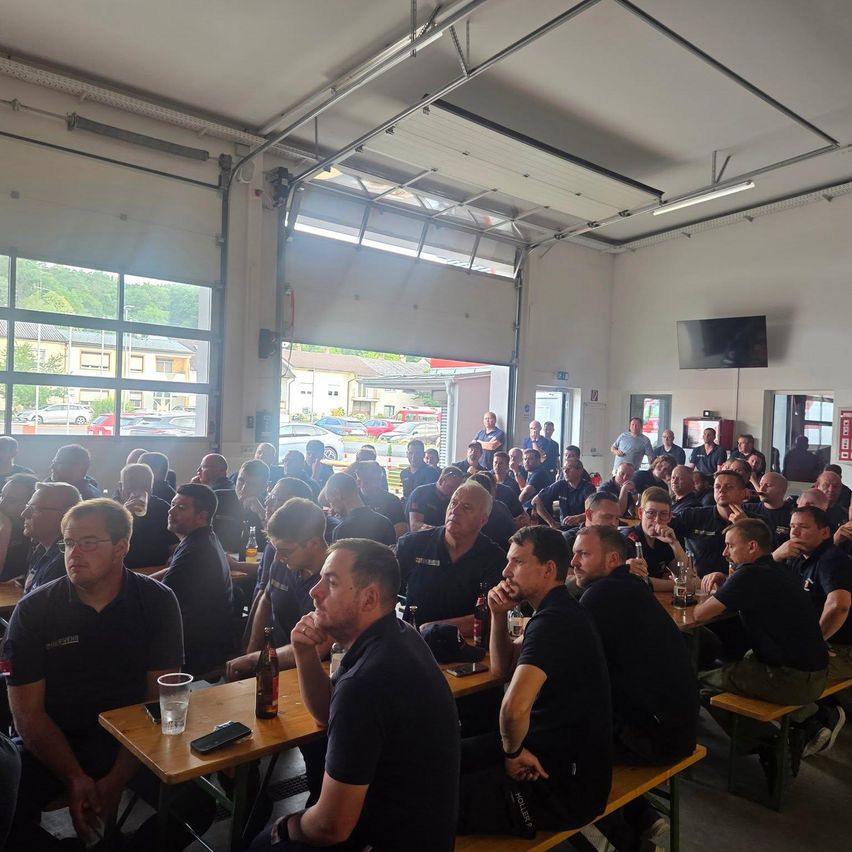 Eine Gruppe von Männern in Uniform sitzt um Tische in einem großen Raum mit großen Fenstern, einige mit Brille. Flaschen und Gläser stehen auf den Tischen, mit einem Monitor an der Wand.