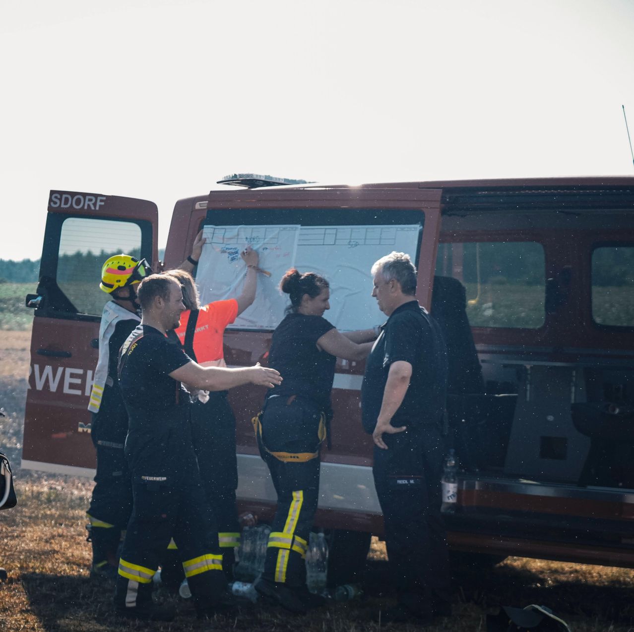 Eine Gruppe von Feuerwehrleuten steht um ein Fahrzeug herum und diskutiert, während eine Frau an eine Tafel schreibt. Sie sind in Uniformen und Helmen gekleidet.