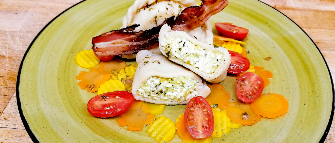 Ein grüner Teller mit drei gefüllten Teigtaschen, Speck, Tomaten und Käse auf einer Holzoberfläche.