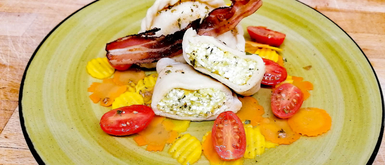 Ein grüner Teller mit drei gefüllten Teigtaschen, Speck, Tomaten und Käse auf einer Holzoberfläche.