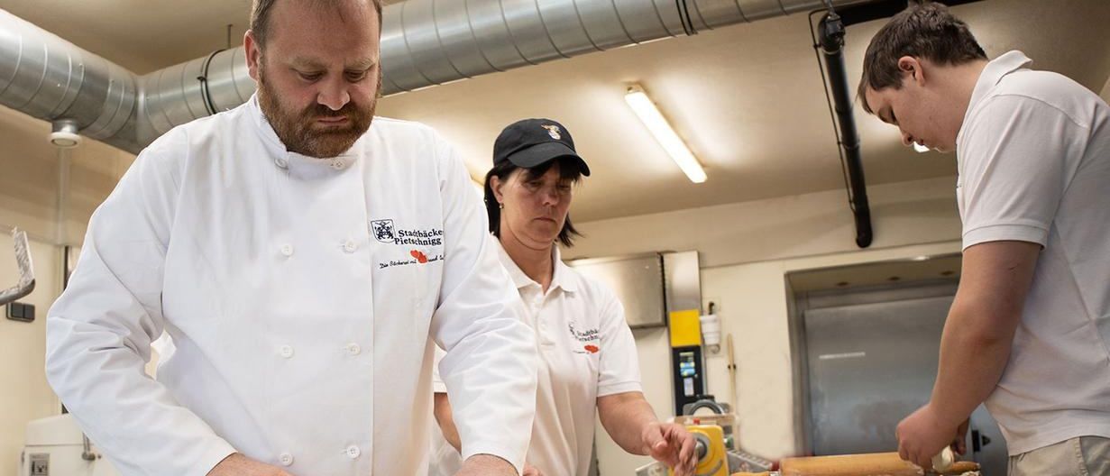 Ein Koch und zwei weitere Personen arbeiten in einer Küche. Der Koch schneidet Teig mit einem Messer. Die anderen bereiten Teig auf einem Tisch vor. Die Küche hat Metallrohre, Lampen und verschiedene Küchengeräte.