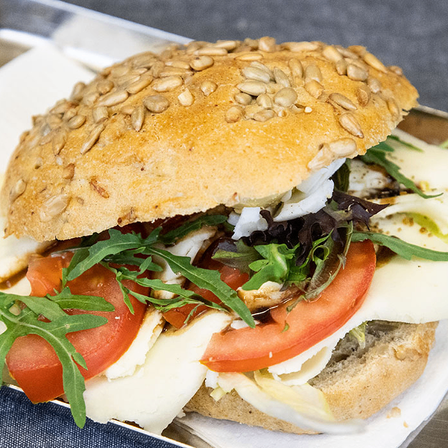 Ein Sandwich mit einem Sesamsamenbrötchen, belegt mit Tomaten, Mozzarella, Salat und Rucola, serviert auf einem silbernen Tablett.