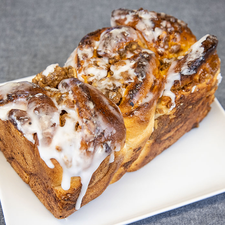Zwei Scheiben Zimtschneckenbrot mit Zuckerguss auf einem weißen Teller. Das Brot ist eine Mischung aus braun und weiß, mit sichtbaren Nüssen und einer knusprigen Kruste.