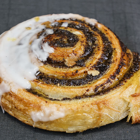 Eine Nahaufnahme eines Zimtschneckens mit weißer Creme, schwarzer Füllung und goldbrauner Kruste auf einer grauen Oberfläche.