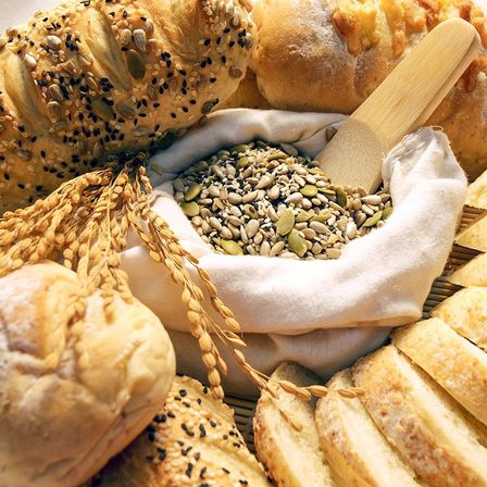 Verschiedene Brotsorten mit Samen und Körnern in einem weißen Beutel, gekrönt von einem Holzspatel, umgeben von Weizenhalmen und Brotscheiben.