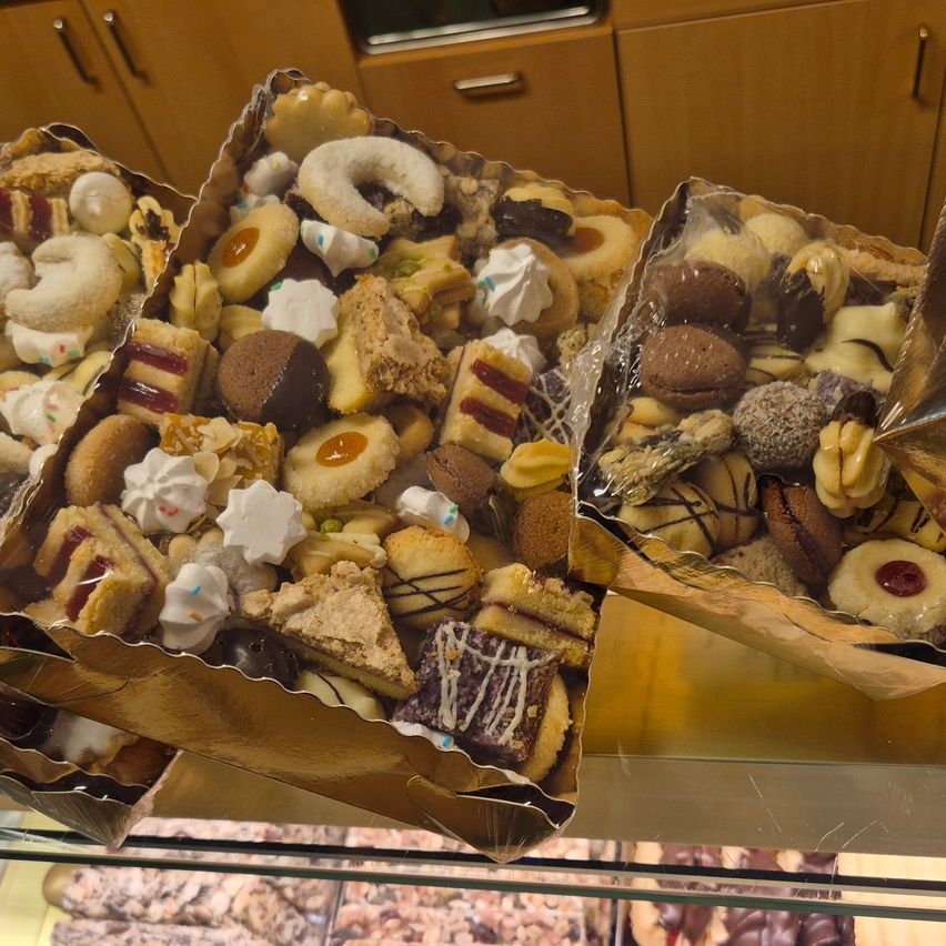 Drei Schachteln mit einer Auswahl an Gebäck, darunter Kekse, Kuchen und Schokoladen, sind auf einem Glasregal ausgestellt. Die Gebäckstücke befinden sich in goldenen Schachteln mit Plastikabdeckungen.