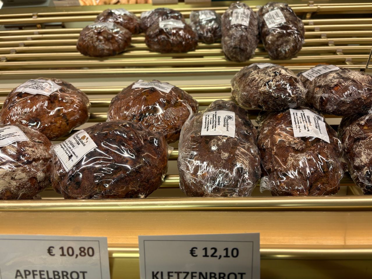 Ein goldener Display-Rack zeigt mehrere Laibe dunklen Brots, eingewickelt in Plastik. Die Preise sind in Euro angegeben. Einer zeigt 10,80 € und der andere 12,10 €.