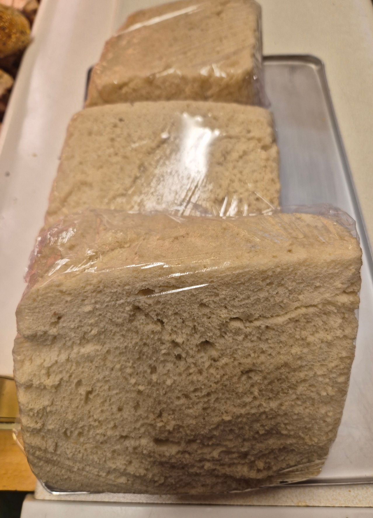 Drei Brote sind ordentlich in einer Reihe angeordnet, jeder in Plastik eingewickelt. Das Brot hat eine goldbraune Kruste und ein weiches, strukturiertes Inneres.