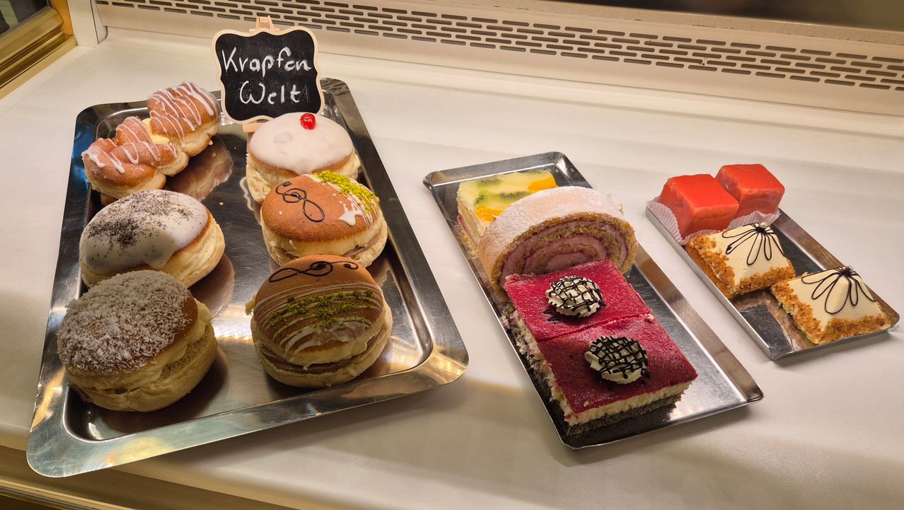 Zwei Tabletts mit Desserts sind ausgestellt. Ein Tablett hat Donuts mit verschiedenen Toppings. Das andere Tablett hat eine Vielfalt von Gebäck, einschließlich eines rot gefüllten Kuchens und einer Spiral-Patisserie.