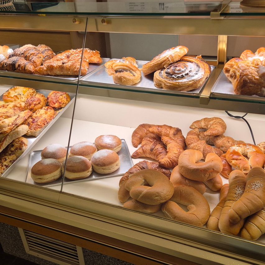 Eine Glasvitrine mit Tabletts gefüllt mit verschiedenen Gebäcksorten, darunter Croissants, Brötchen und Brezeln, auf drei Ebenen angeordnet.
