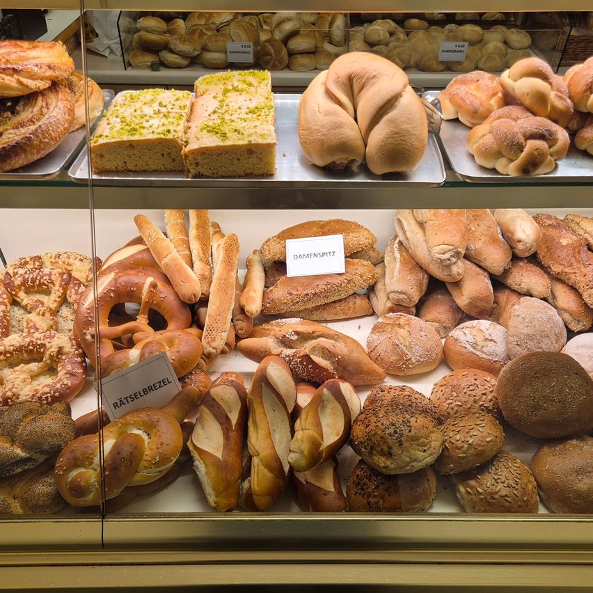 Eine Glasvitrine gefüllt mit verschiedenen Broten und Gebäck, darunter Brezeln und Brötchen mit Toppings.