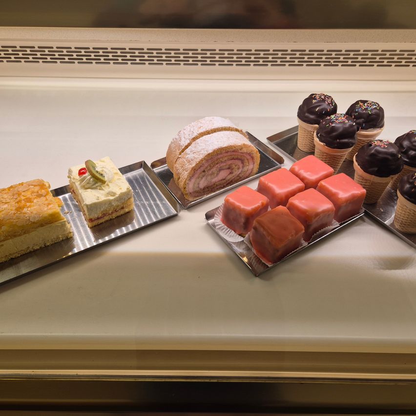 Verschiedene Desserts auf Tabletts ausgestellt, darunter ein Stück Kuchen, eine Schokoladenrolle und rote quadratische Bonbons. Schokoladenkegel mit Streuseln sind ebenfalls vorhanden.