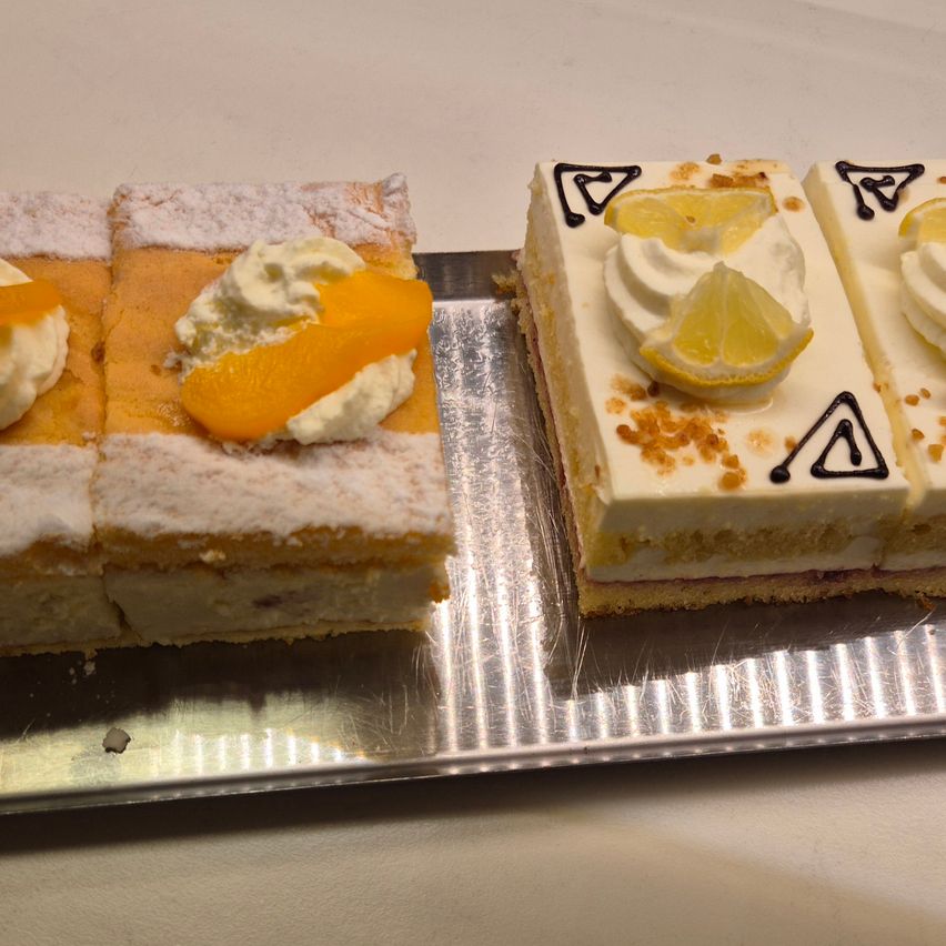 Zwei Desserts stehen auf einem Silbertablett. Eines hat Aprikosenscheiben und Sahne, das andere hat Zitronenscheiben und schwarze Schwünge. Beide haben eine Schicht Biskuit und weiße Glasur.