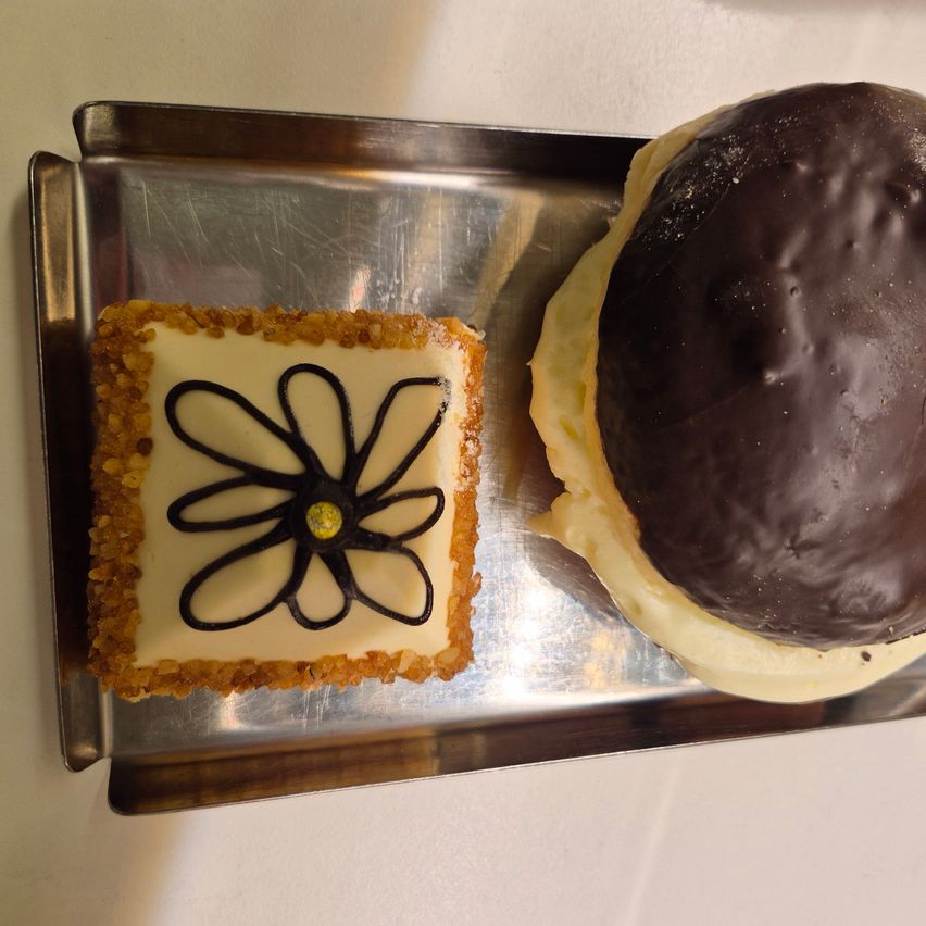 Zwei Desserts auf einem silbernen Tablett. Eines ist ein Quadrat mit Blumendesign und gelbem Zentrum, das andere ist ein runder, schokoladenüberzogener Kuchen.