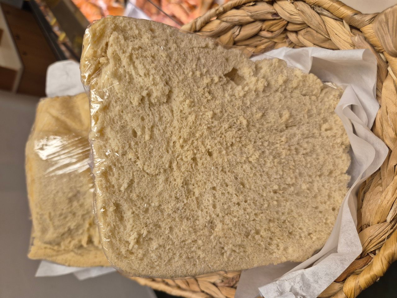 Eine Nahaufnahme eines Brotlaibs, teilweise geschnitten, in Plastik eingewickelt und in einem geflochtenen Korb platziert. Ein weiterer Laib ist teilweise links sichtbar.