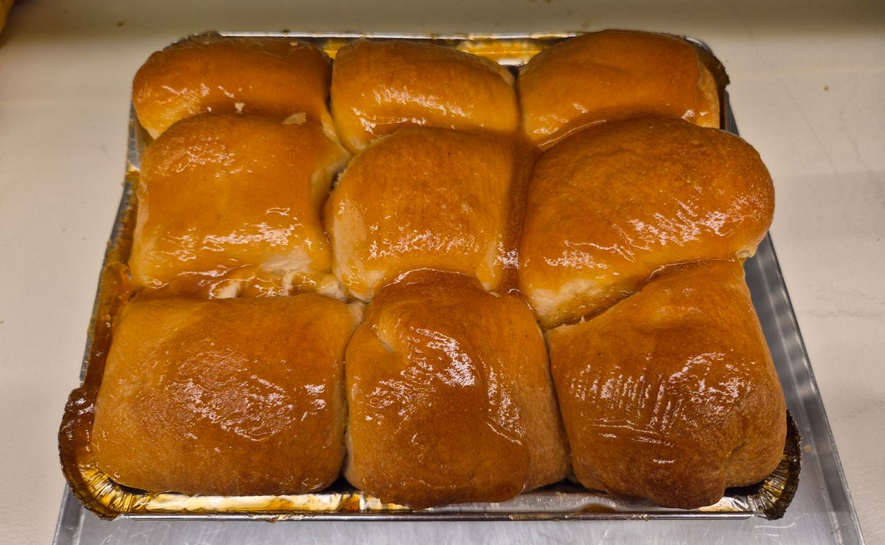 Eine Schale mit goldbraunen, glasierten Donuts, leicht zusammengedrückt, mit einer glänzenden Oberfläche. Die Schale ist metallisch und auf einem weißen Hintergrund platziert.