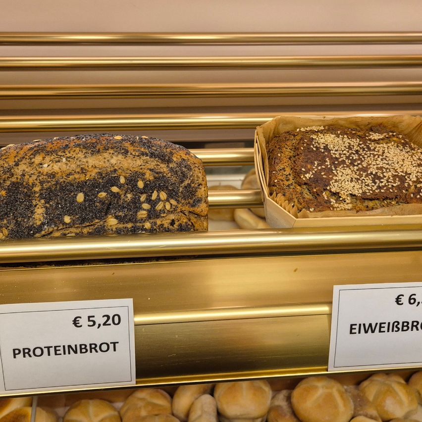 Zwei Brotsorten auf einem goldenen Regal. Eines ist mit 'Proteinbrot' beschriftet und kostet 5,20 €, das andere ist mit 'Eiweißbrot' beschriftet und kostet 6 €.