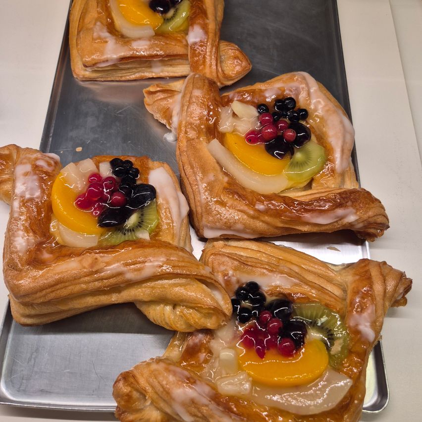 Fünf blättrige Gebäckstücke mit Fruchtfüllungen, darunter Kiwi, Beeren und Orangenscheiben, sind auf einem Metalltablett angeordnet. Die Gebäckstücke sind glasiert und haben eine goldbraune Farbe.