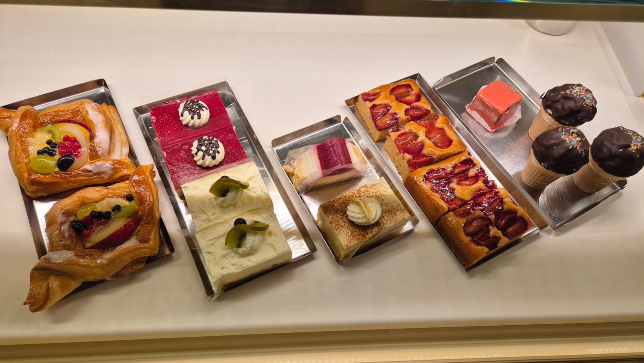 Fünf Tabletts mit Desserts sind auf einer weißen Oberfläche ausgestellt. Jedes Tablett enthält eine Vielzahl von Kuchen und Gebäck, darunter Red Velvet Cake, Käsekuchen und mit Früchten garnierte Desserts.