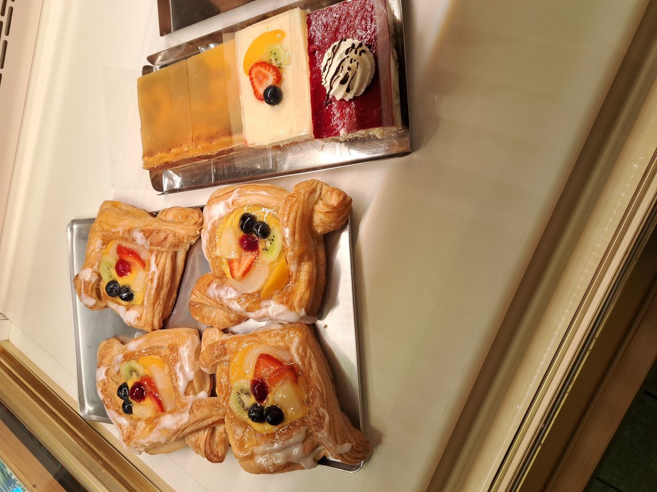 Ein Tablett mit Gebäck, darunter Obst-gefüllte Croissants und quadratische Desserts mit Obstscheiben und Schlagsahne. Das Gebäck steht auf einer weißen Arbeitsplatte.