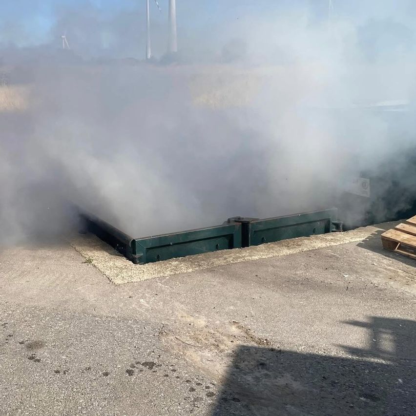 Ein grauer Betonbereich mit Rauch, der aus ihm kommt, befindet sich im Freien. Eine grüne Metallbarriere steht in der Mitte. Rechts ist ein Holzpalette.
