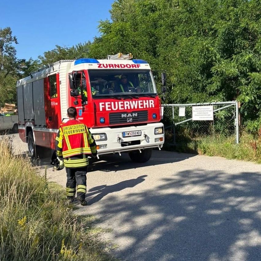 Ein Feuerwehrmann geht auf einen roten Feuerwehrwagen zu, der mit 'Feuerwehr' beschriftet ist. Der Wagen steht auf einer Straße, und in der Nähe befindet sich ein Metallzaun mit Schildern.