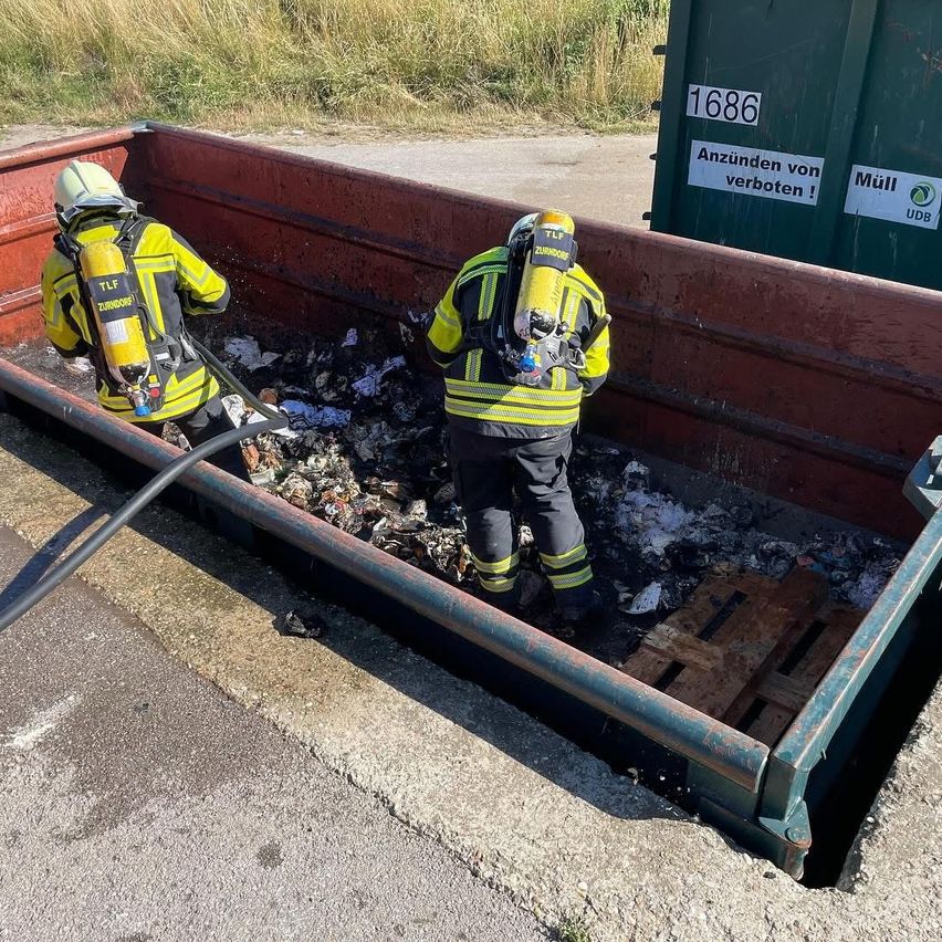 Zwei Feuerwehrleute in gelben Uniformen und Helmen räumen einen Müllcontainer mit Müll und Schutt auf. Dahinter steht ein grüner Container mit einem Schild, das 'Anzunden von verboten!' besagt.